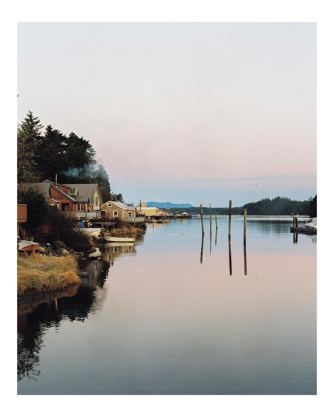 A small town with houses and boats along a calm body of water is visible under a soft, colorful sky.