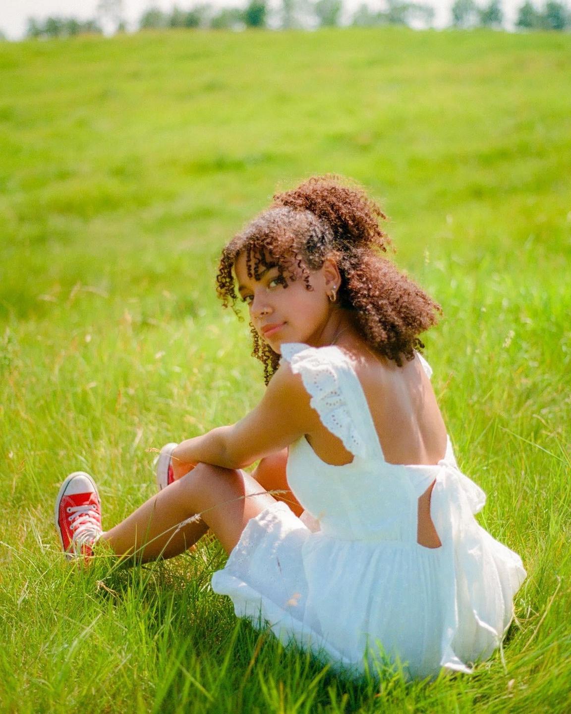 A young woman with curly hair sits in a field of green grass, looking over her shoulder at the viewer.