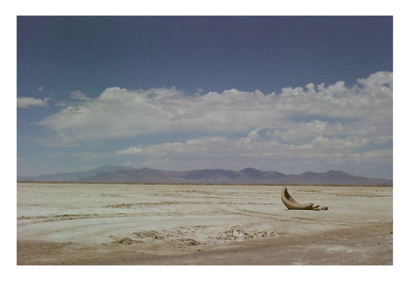A large, curved, light-colored object lies on a vast, flat, dry landscape under a partly cloudy sky with mountains in the distance.