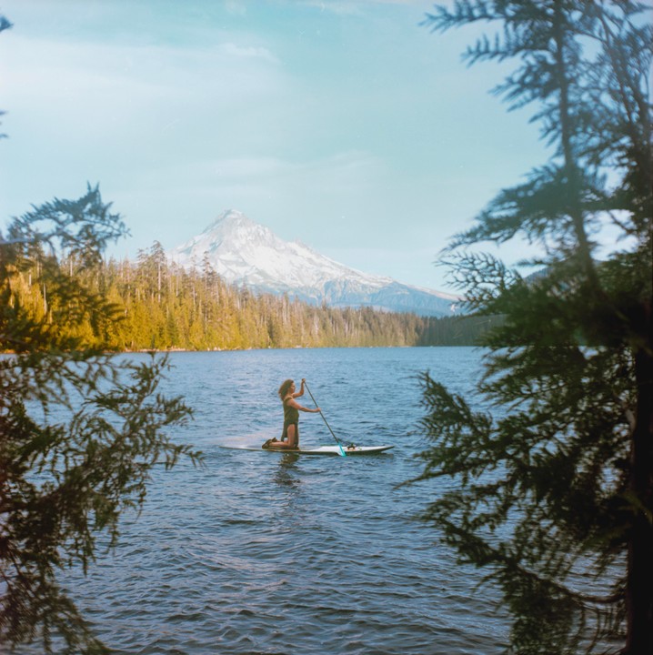A woman kneels on a paddleboard in a lake with a snow-capped mountain and forest in the background.