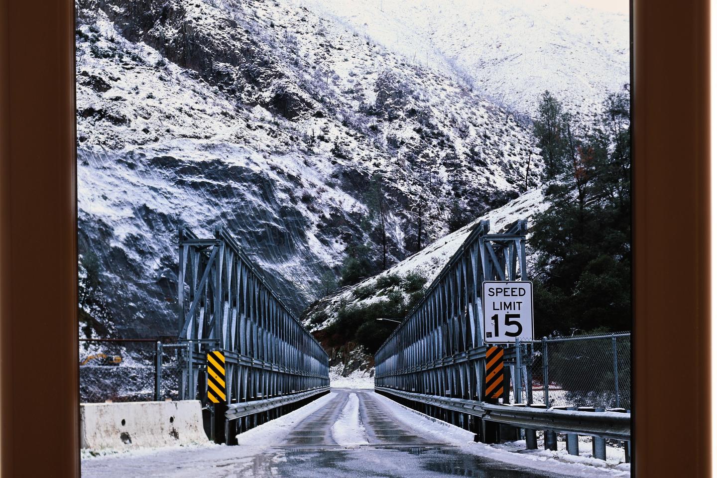 A snowy mountain road leads to a metal bridge with a speed limit sign.