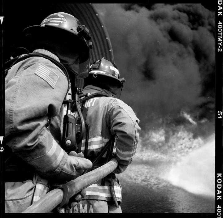Two firefighters in uniform hold a hose and spray water at a large plume of smoke.