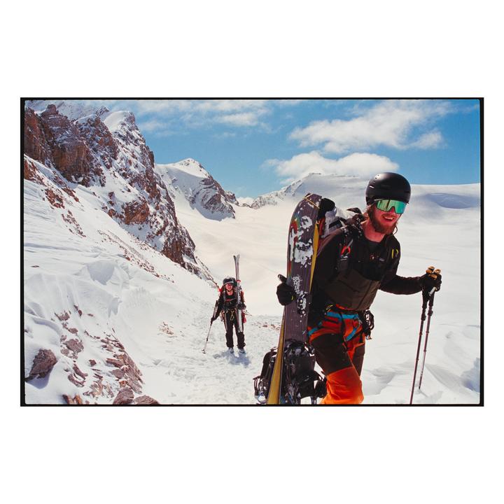 Two people are hiking up a snowy mountain path with skis and snowboards strapped to their backpacks.
