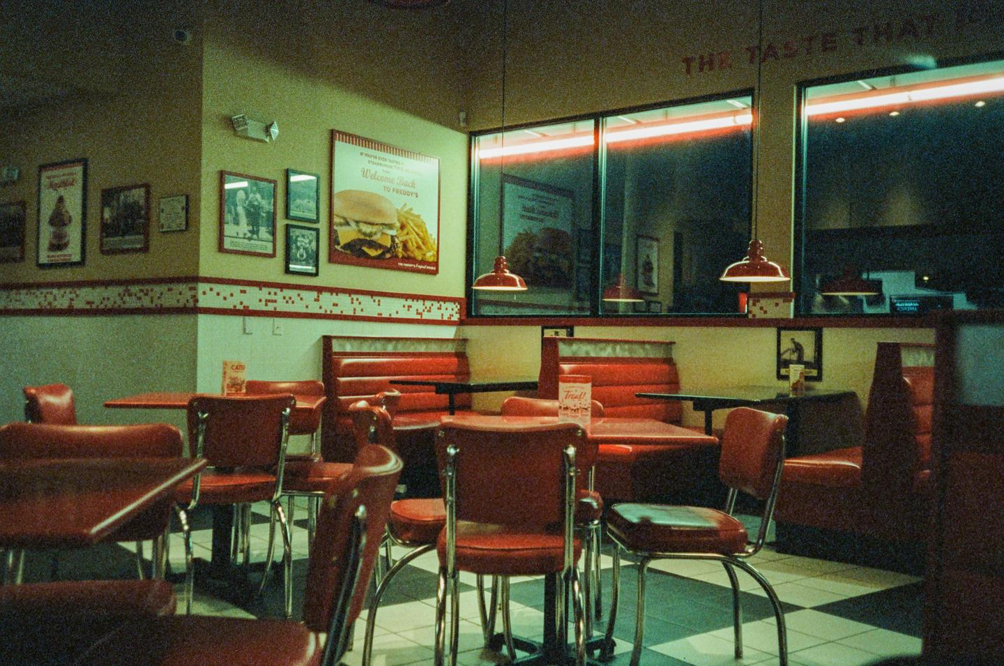 A diner interior with red booths and chairs, checkered floor, and a large burger advertisement on the wall.