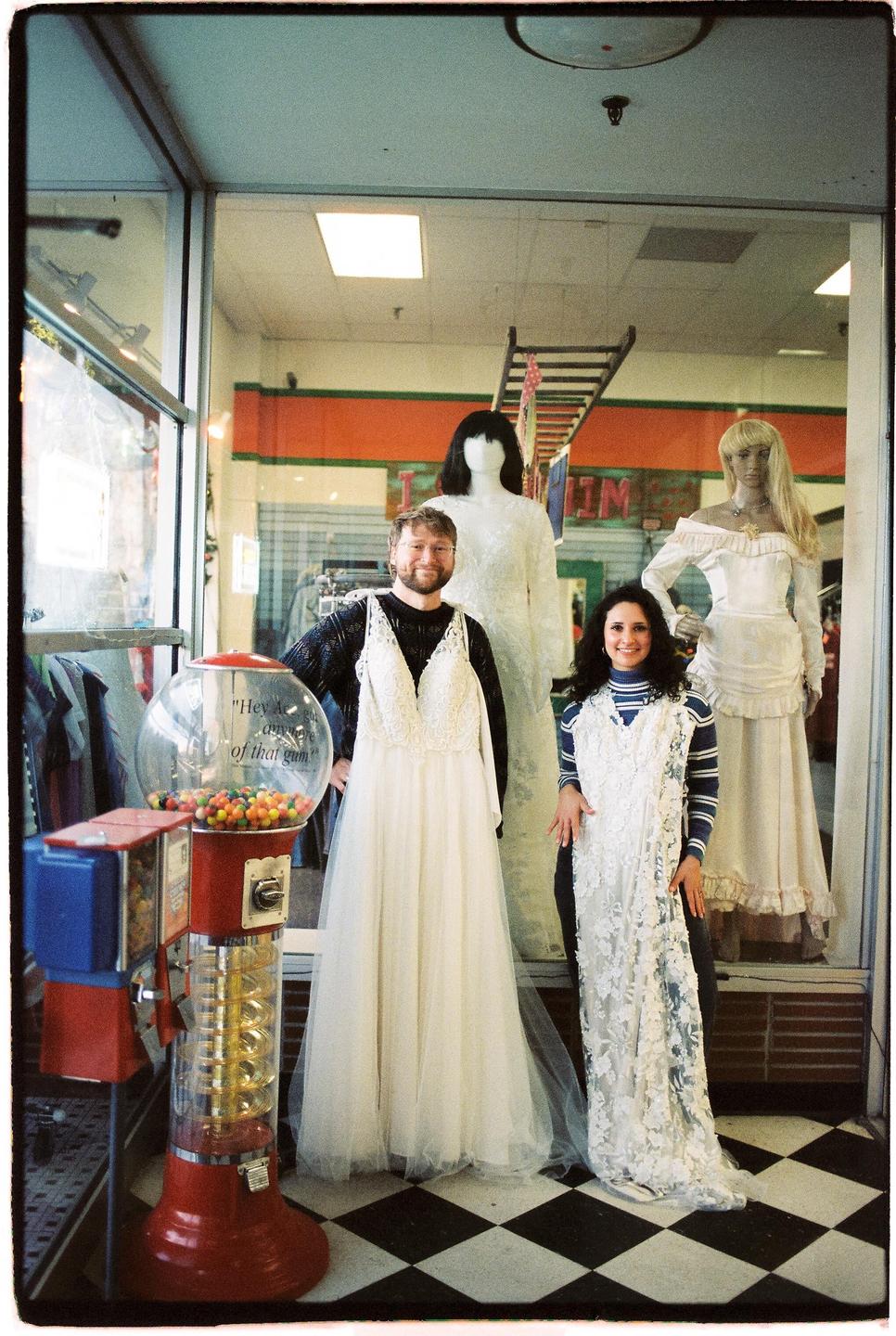 A couple poses with wedding dresses in a store window next to a gumball machine.