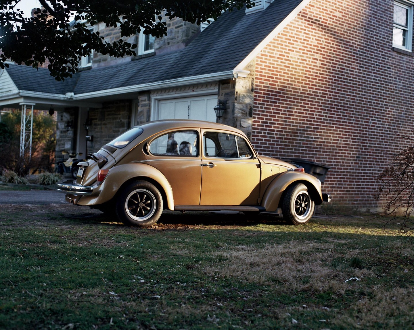 A gold volkswagen beetle is parked on a grassy lawn in front of a house with a brick wall.