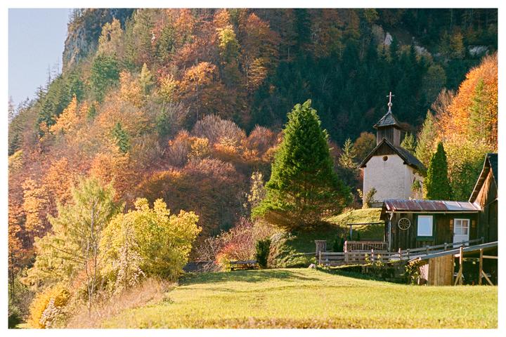 A small chapel and a wooden cabin are nestled on a grassy hill surrounded by a forest with autumn foliage.