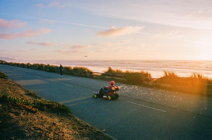 A child in a toy car blows bubbles on a road next to a beach at sunset.