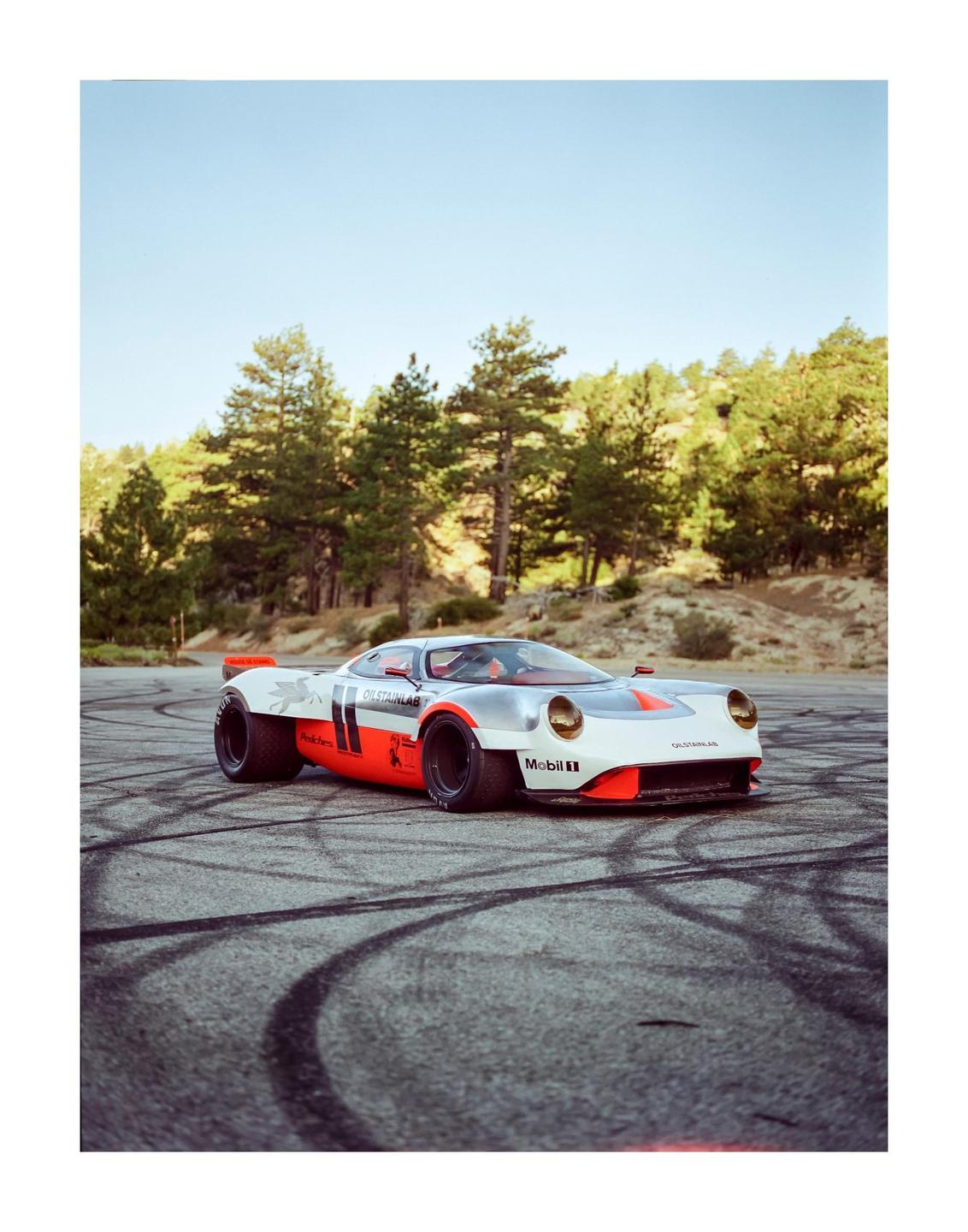 A silver and orange race car with wide tires is parked on an asphalt road with tire marks, surrounded by pine trees and hills.