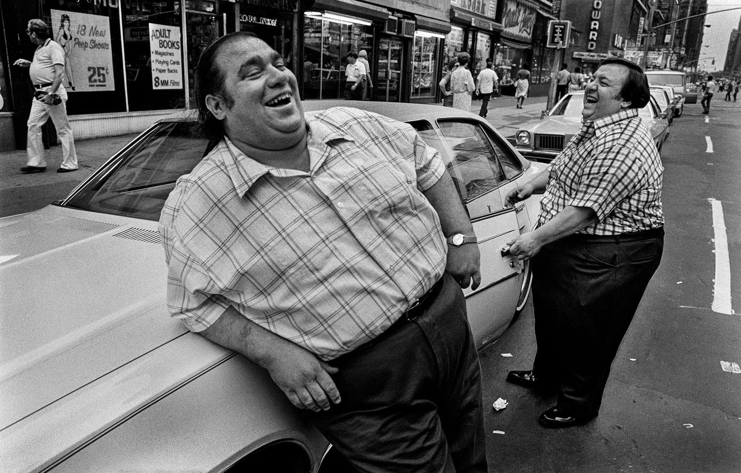 Two men are laughing on a city street next to a car.