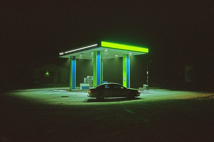 A car is parked at a gas station at night, illuminated by neon lights.