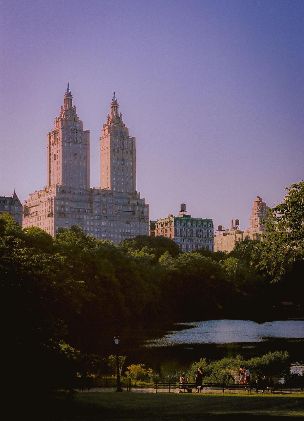 Two tall buildings with ornate spires rise above a treeline, overlooking a body of water where people are gathered on benches.