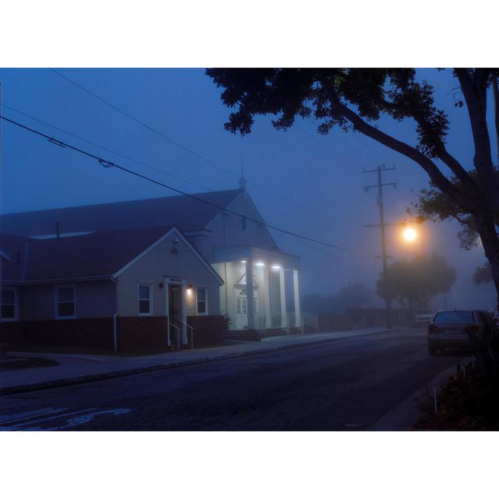 A street light illuminates a foggy street with a church and a house on the left and a car parked on the right.
