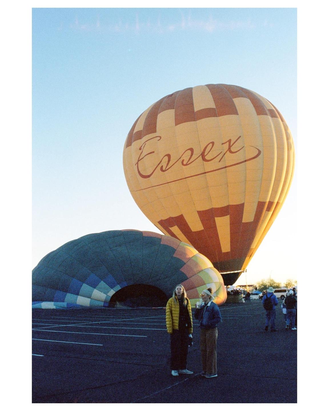 Two women stand in a parking lot next to two hot air balloons, one of which is partially deflated.