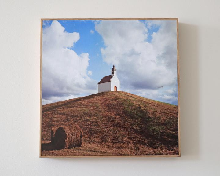 A small white church with a brown roof stands atop a grassy hill under a cloudy sky, with a hay bale in the foreground.