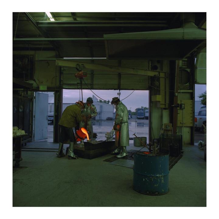 Three men in protective gear pour molten metal into a mold inside a foundry.