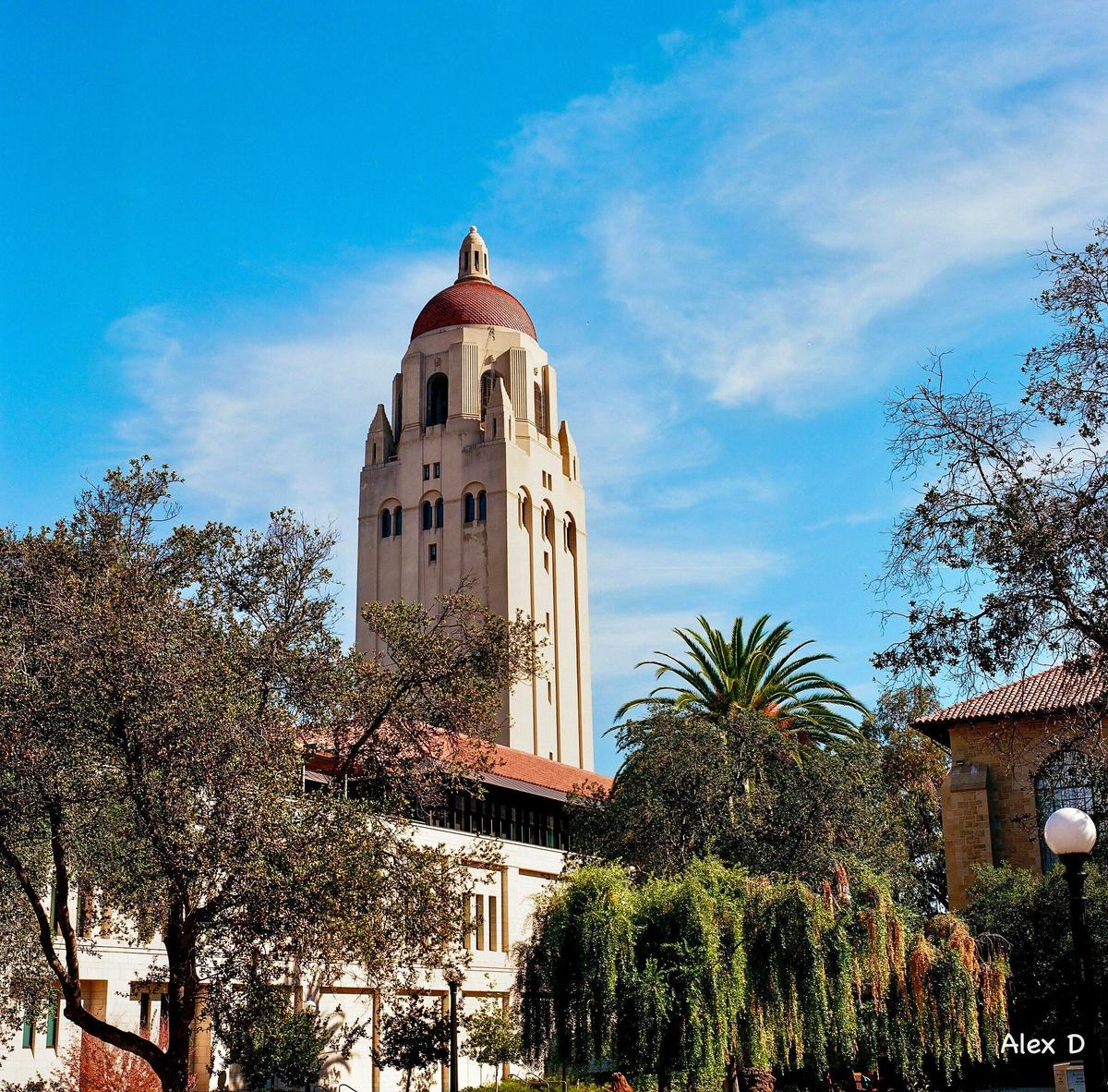 A tall, light-colored tower with a red dome rises above trees and buildings under a blue sky.