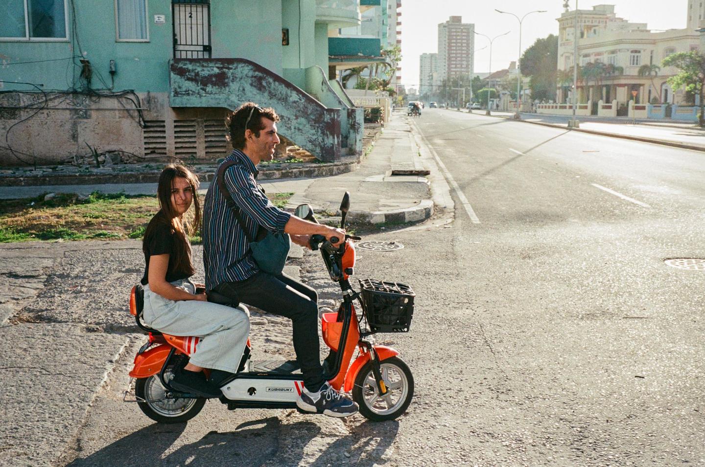 A man and a woman are riding an orange scooter on a street in Havana.