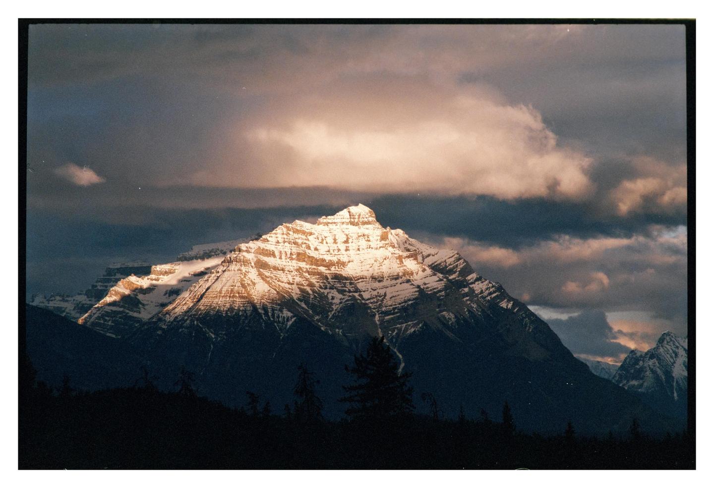 A snow-capped mountain peak is illuminated by a ray of sunlight under a cloudy sky.