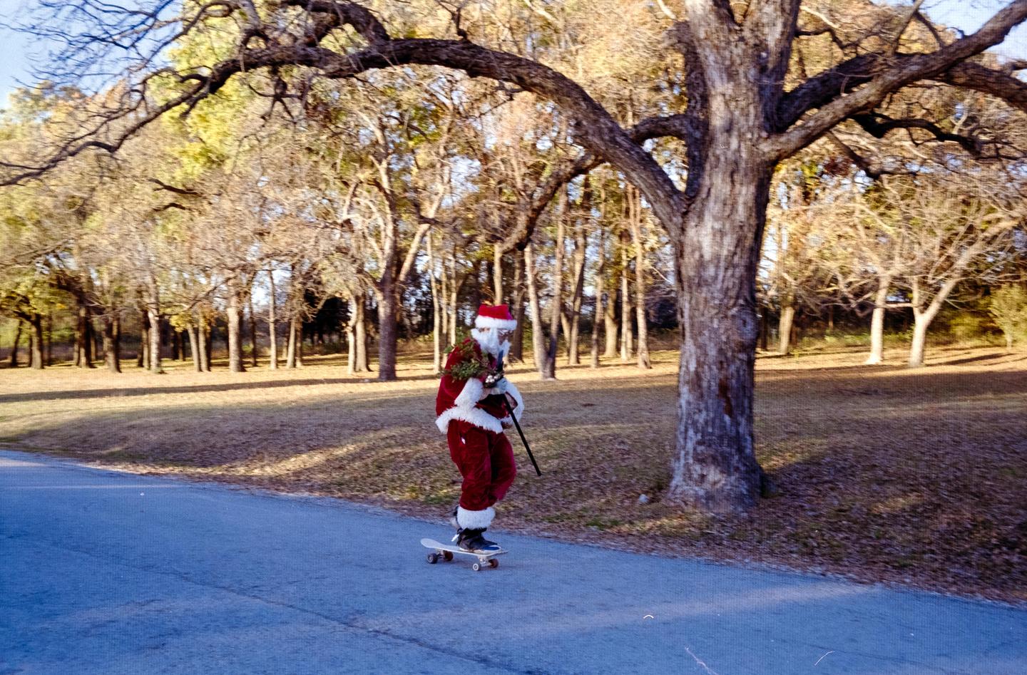 A person dressed as Santa Claus is skateboarding on a paved path next to a grassy area with trees.