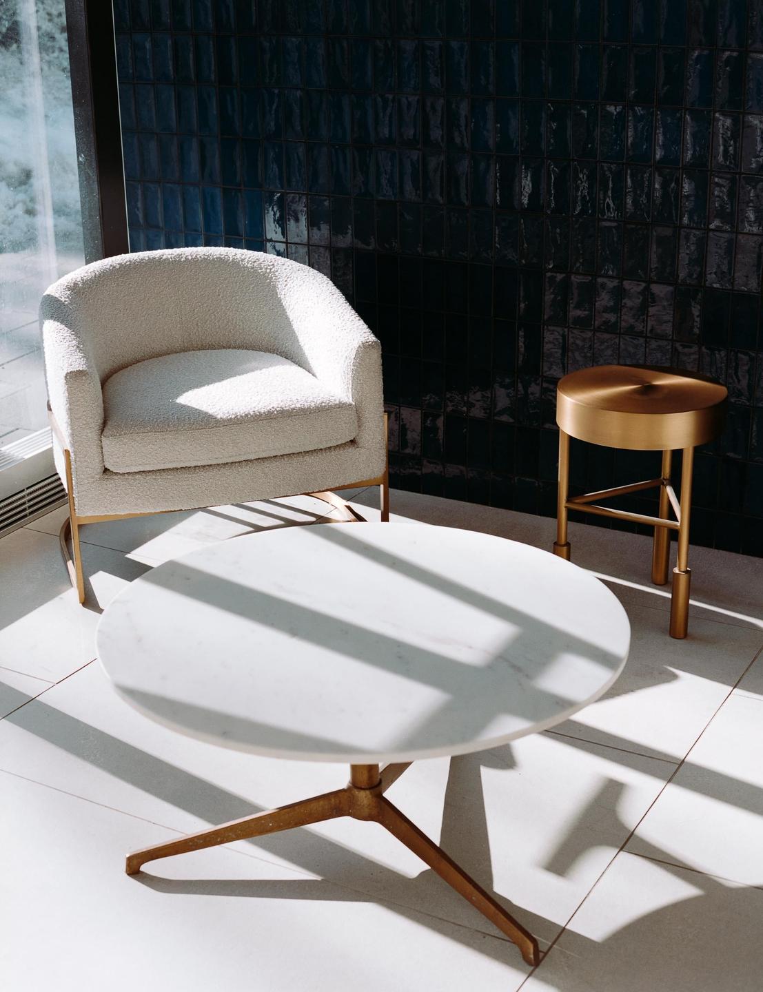 A white armchair, a marble coffee table, and a brass side table are arranged in a room with dark tiled walls and bright sunlight.