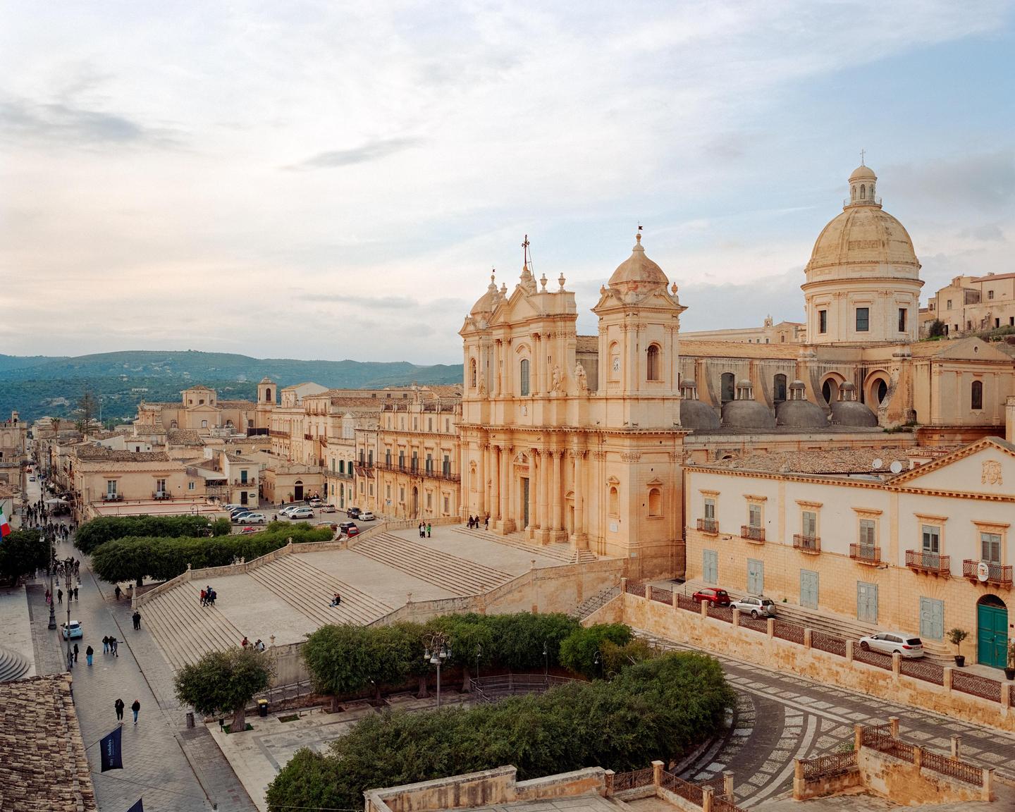 A large baroque church with a grand staircase dominates a town square with people and cars.