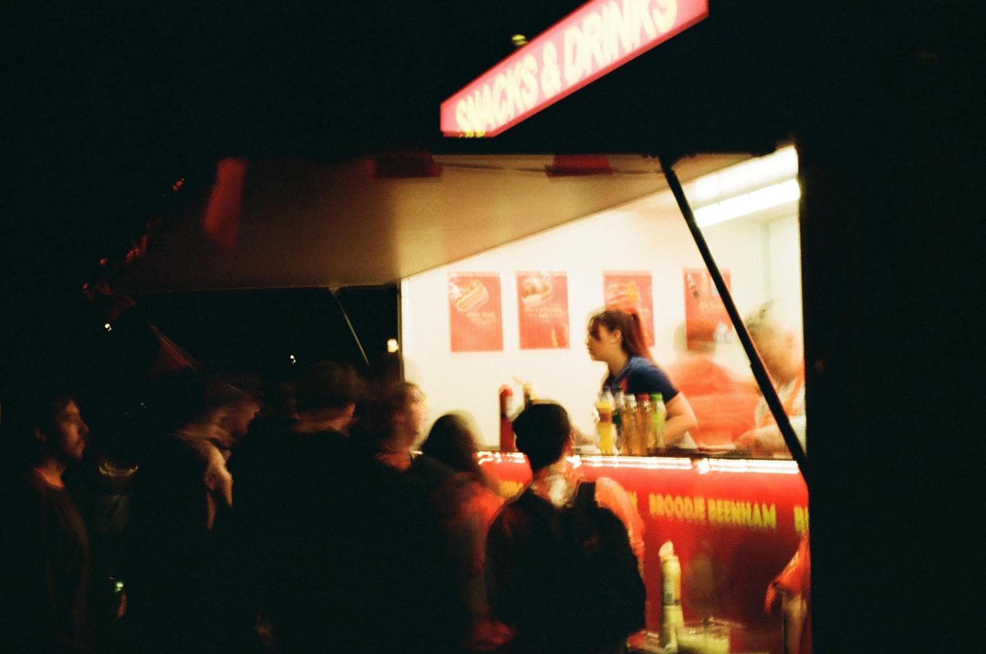 A woman works at a brightly lit food stall at night while customers wait in line.