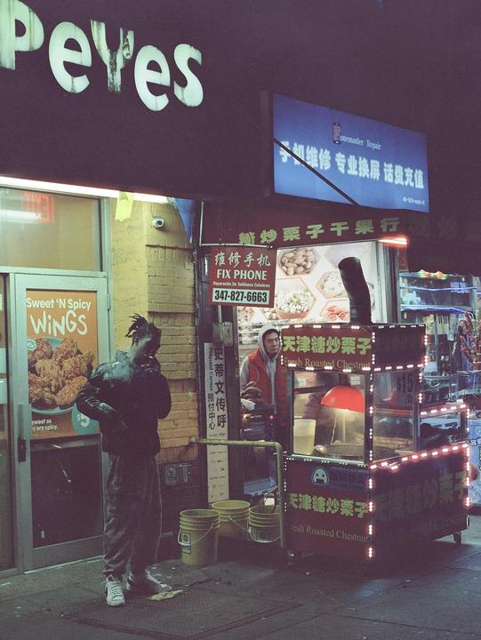 A man stands on a street at night next to a food cart and a fried chicken restaurant.