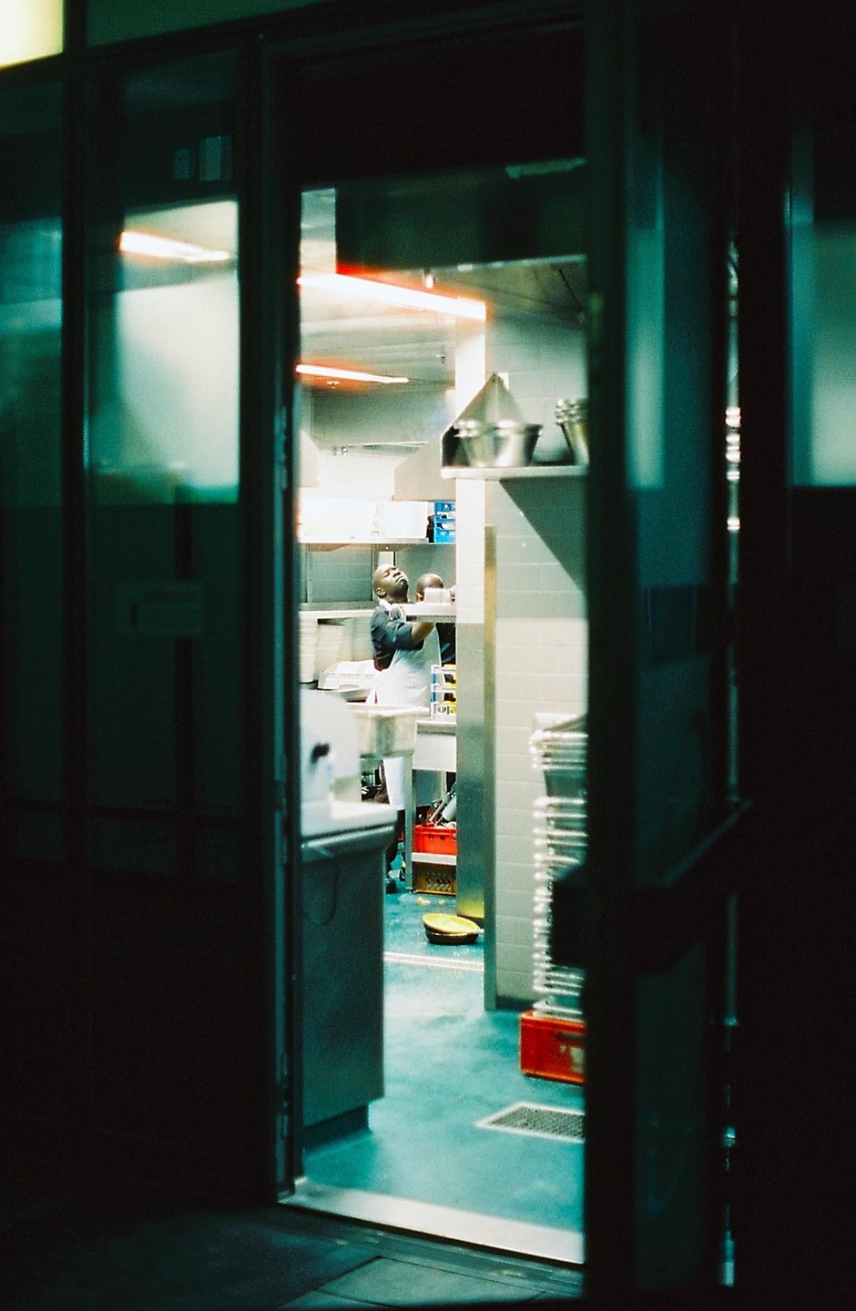 A man in an apron works in a brightly lit kitchen, viewed through a doorway.