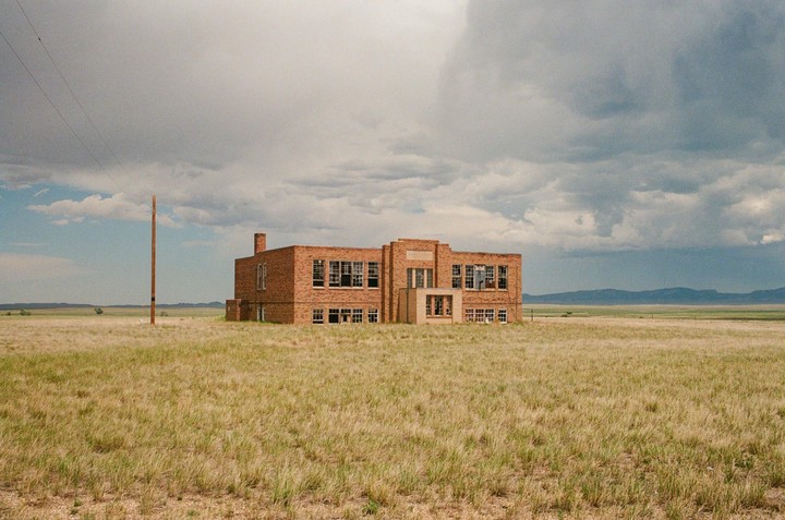 An abandoned brick building stands in a vast, dry grassy field under a cloudy sky, with mountains in the distance.