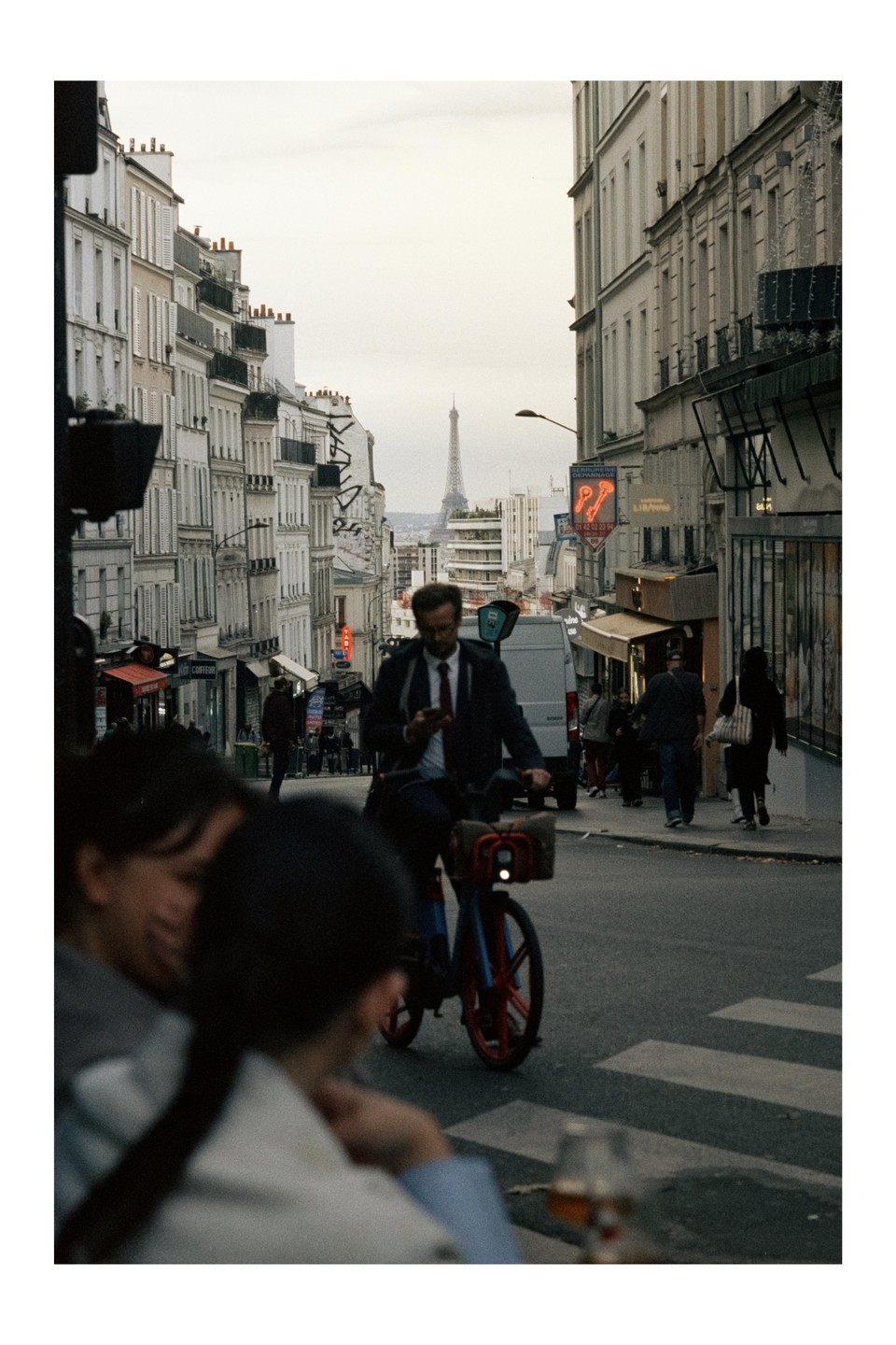 A man on a bicycle looks at his phone while riding down a street with the Eiffel Tower visible in the distance.
