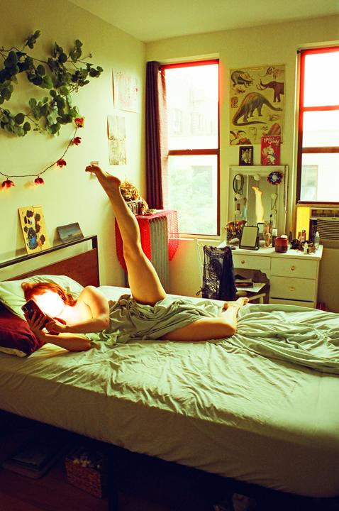 A woman lies on a bed with one leg raised, looking at her phone in a sunlit room.