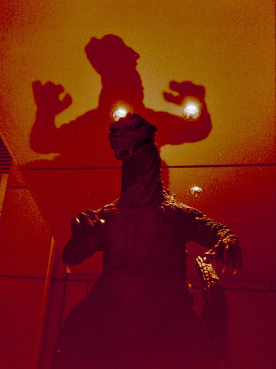 A Godzilla statue casts a large shadow on a red and orange wall.