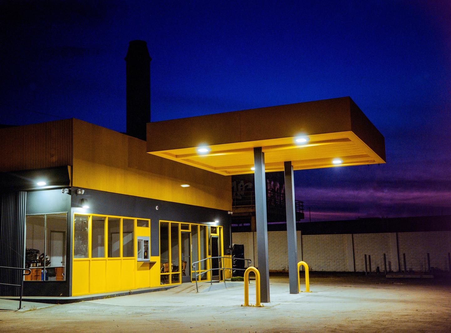A gas station with yellow accents is illuminated at night under a deep blue and purple sky.