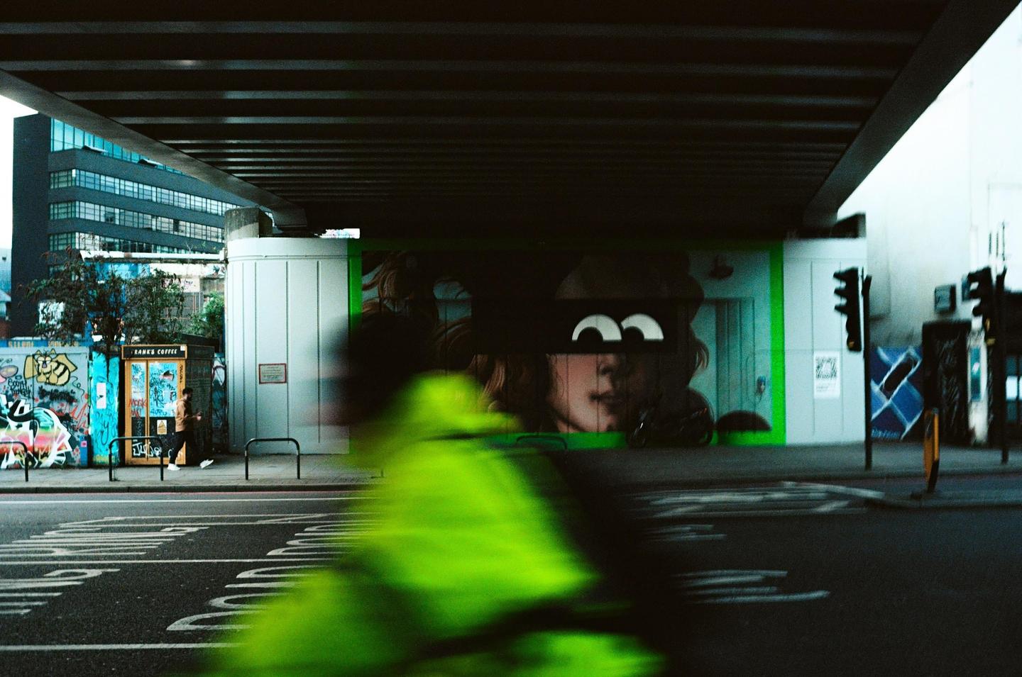 A blurred green shape moves past a street mural of a woman's face under an overpass.