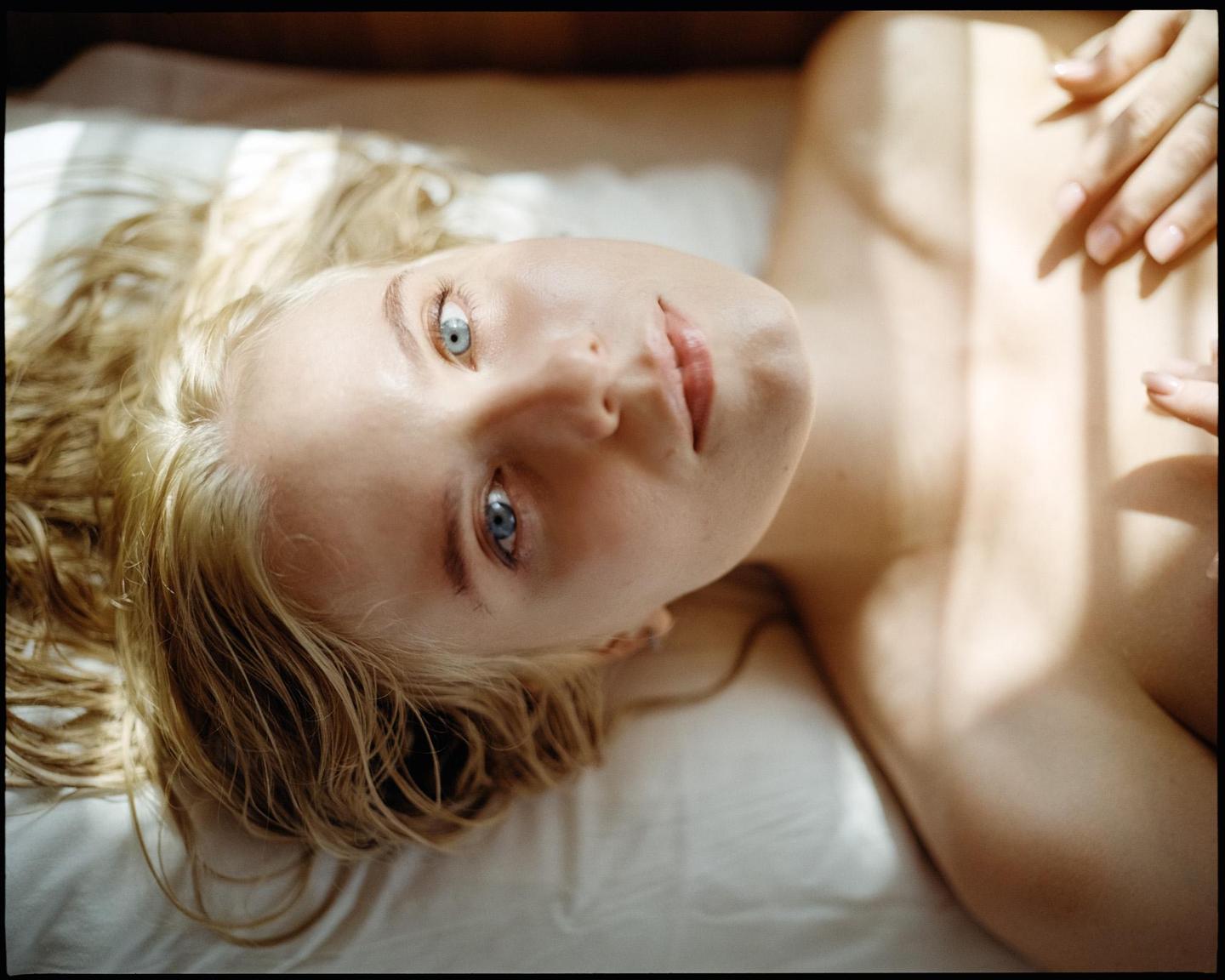 A woman with blonde hair and blue eyes lies on a white sheet, looking up at the viewer.