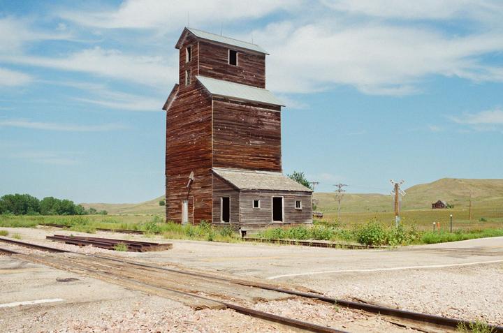 An abandoned wooden grain elevator stands beside railroad tracks in a rural landscape under a blue sky with clouds.