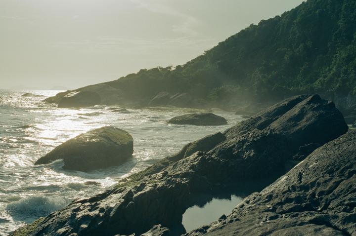 Waves crash against rocks on a hazy beach with a lush green hill in the background.