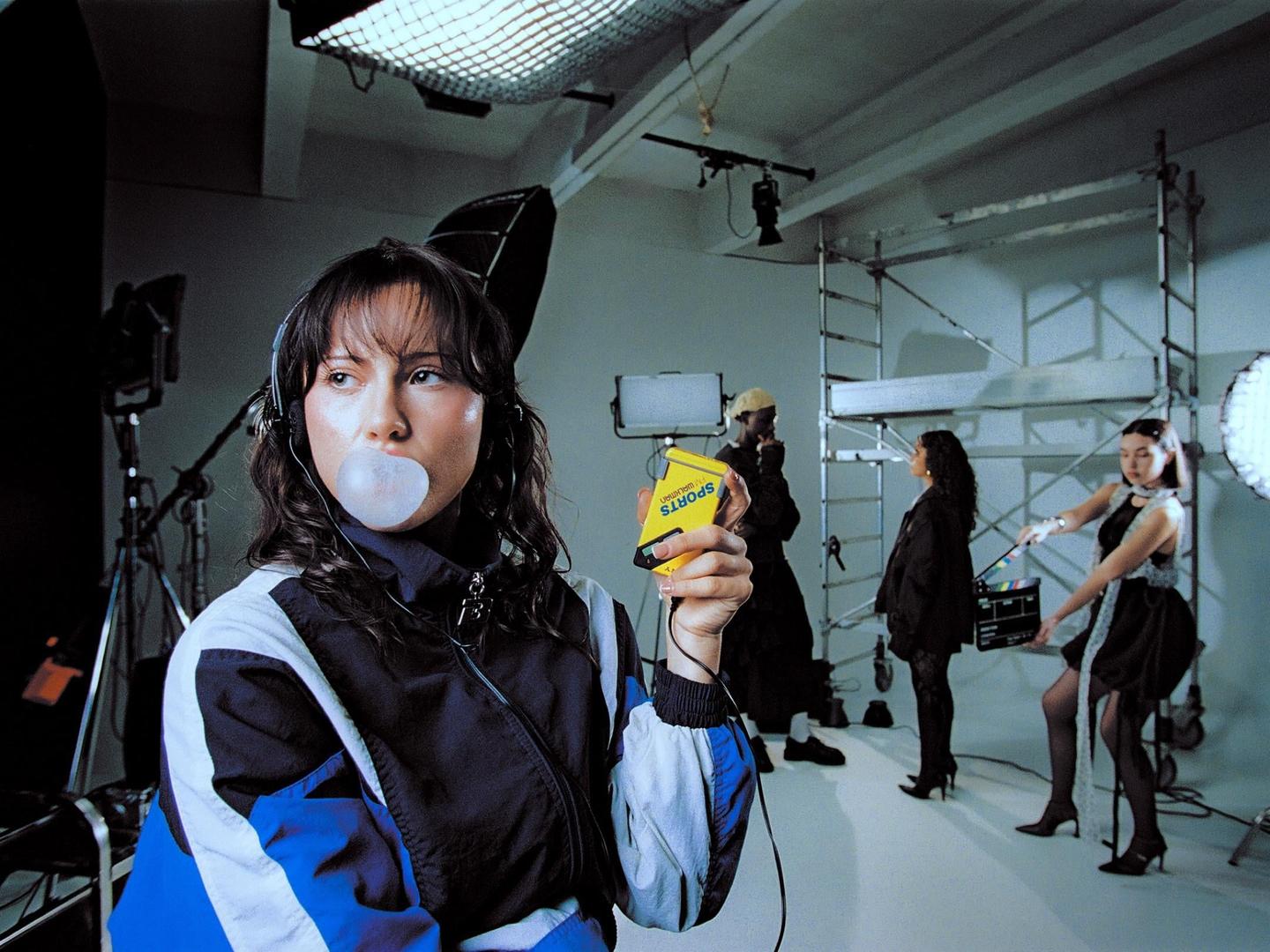 A woman with headphones blows a bubblegum bubble while holding a yellow device in a studio with other people and equipment.