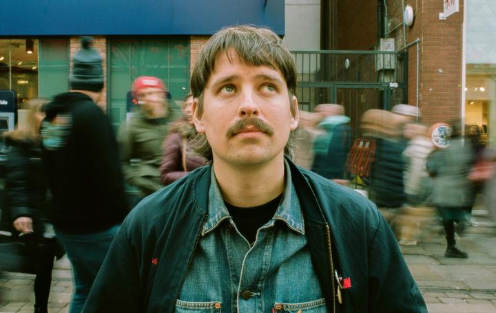 A man with a mustache and mullet looks up while standing on a street with blurred people walking in the background.