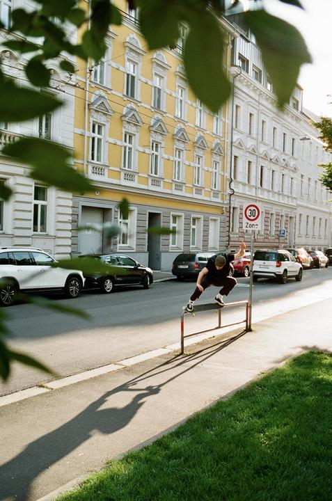A man on a skateboard grinds a metal barrier on a city street lined with parked cars and buildings.