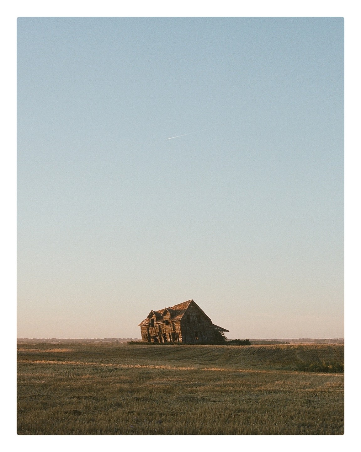 An old wooden house stands in a field under a clear sky with a contrail.