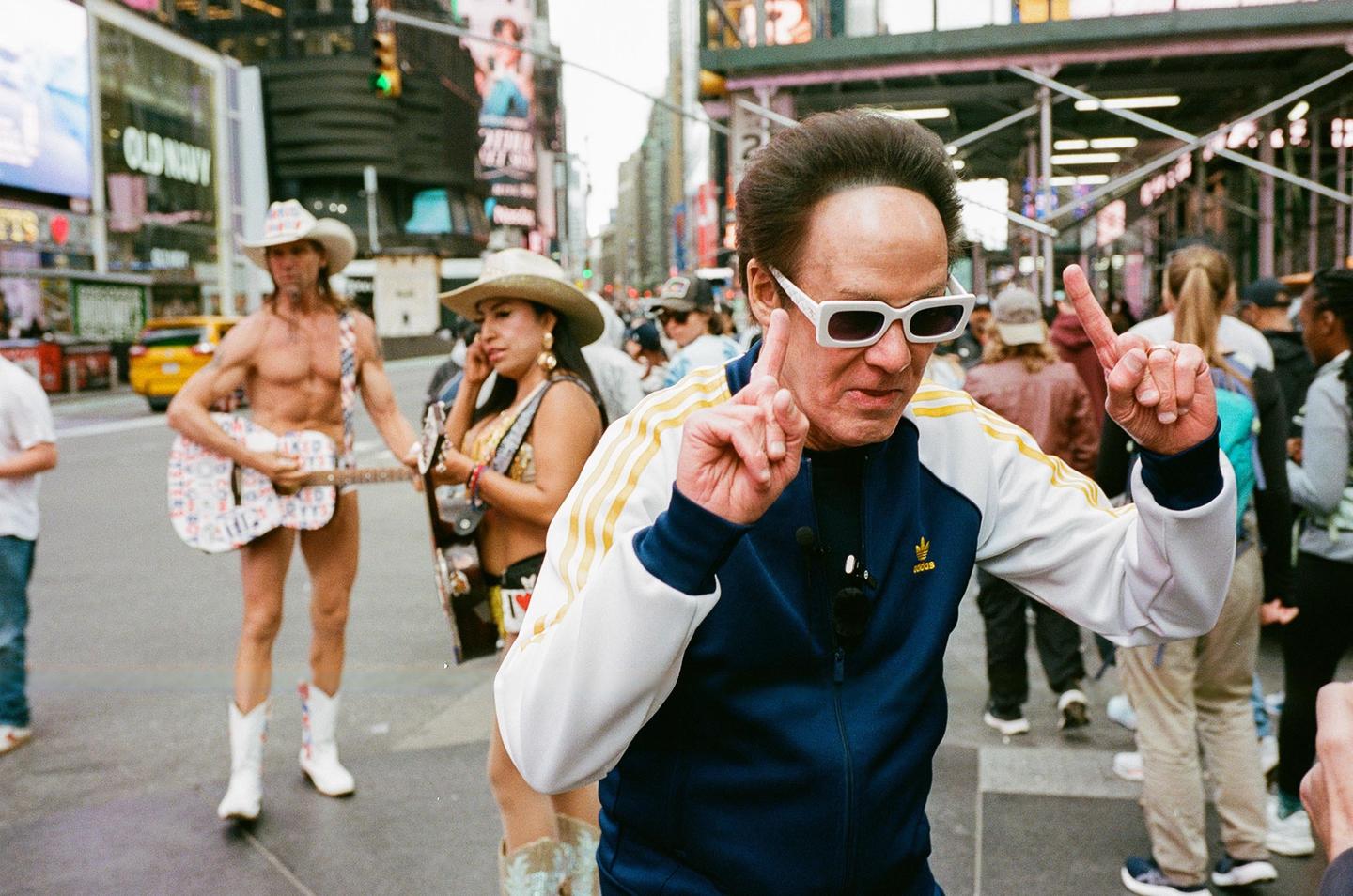 A man in a tracksuit and white sunglasses points his fingers up while a man in a cowboy hat and underwear plays a guitar and a woman in a cowboy hat stands nearby on a city street.