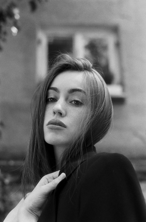 A woman with long hair and freckles looks directly at the viewer, with a hand on her shoulder.