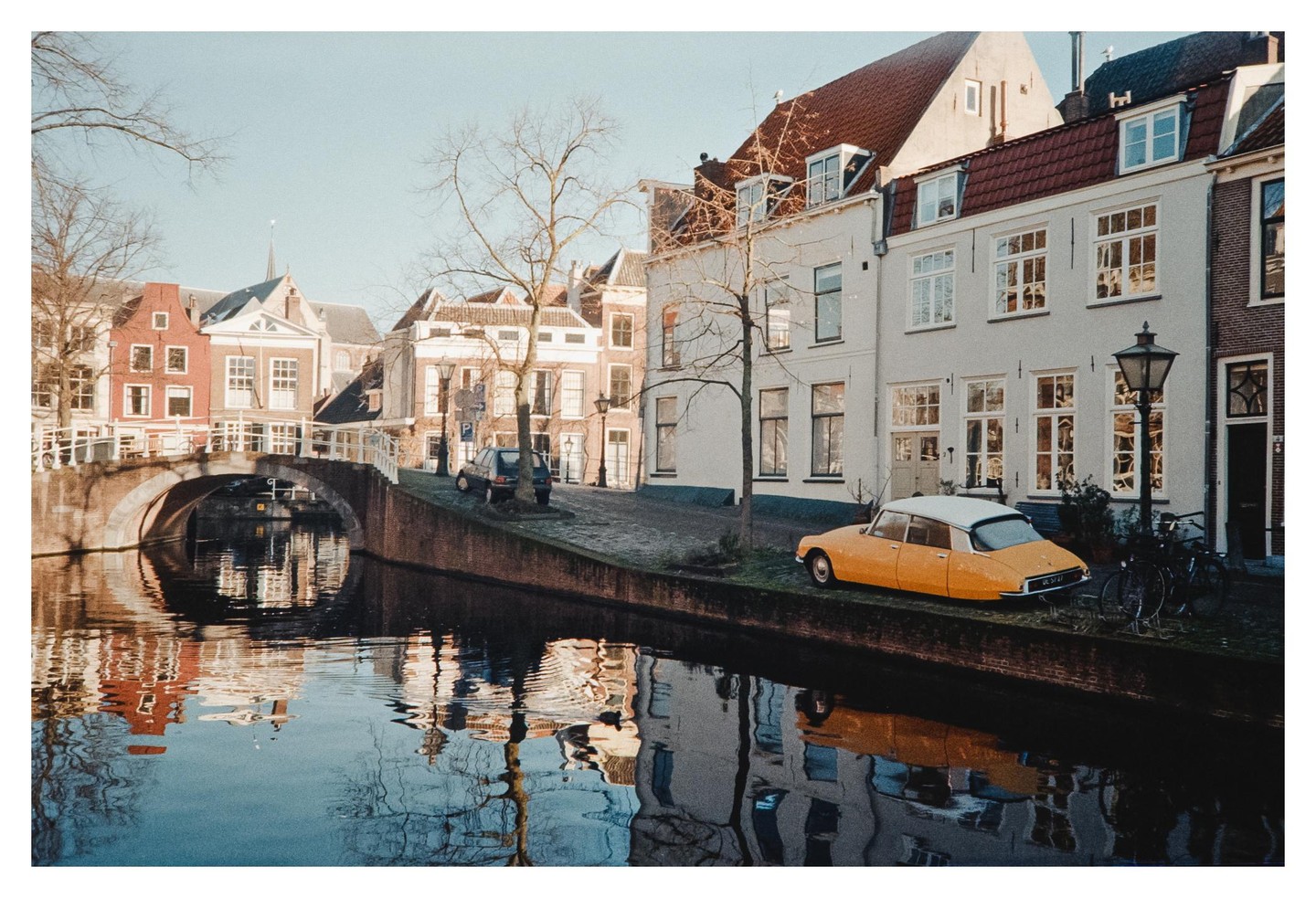 A yellow car is parked on a cobblestone street next to a canal with buildings and a bridge in the background.