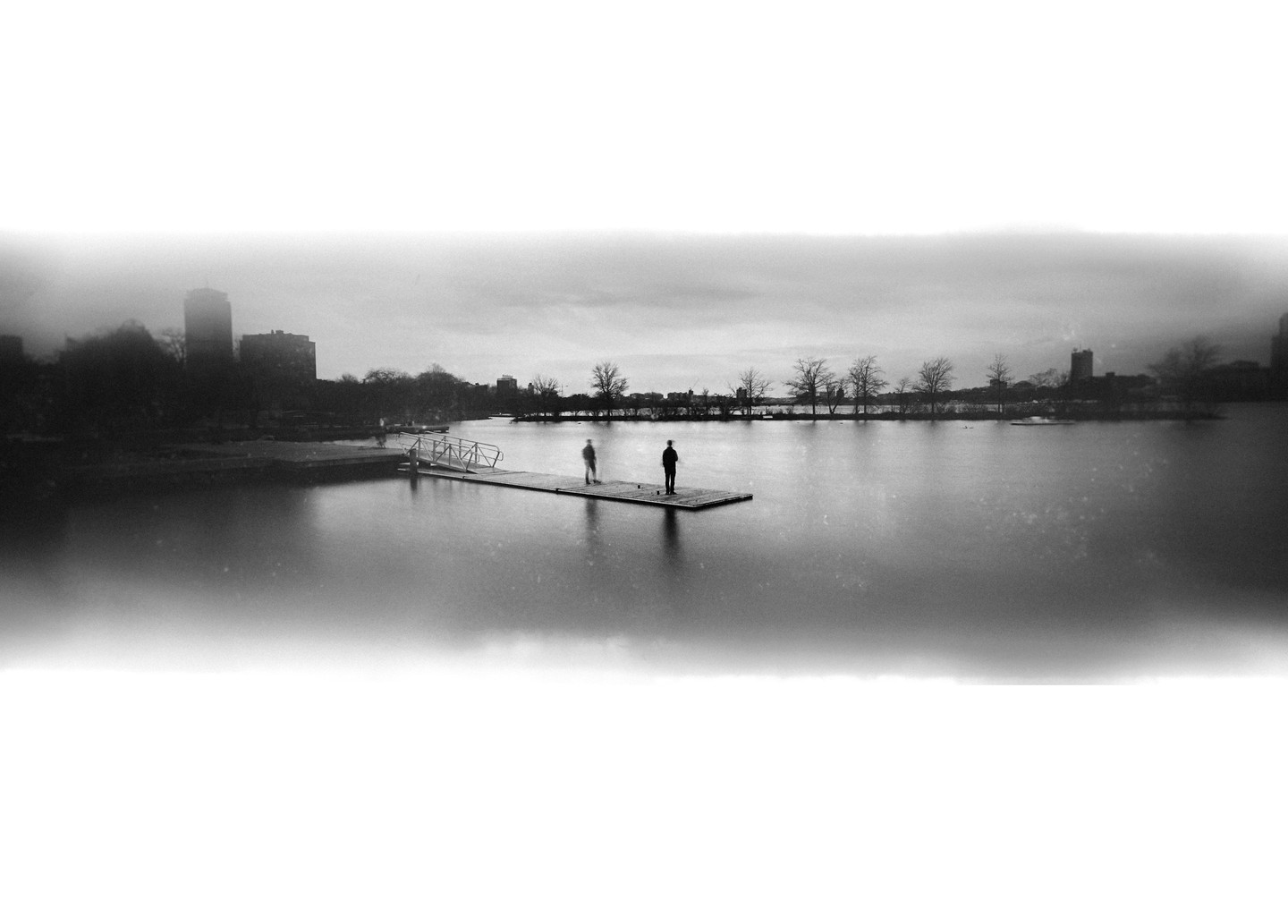 Two men stand on a dock extending into a wide body of water with a city skyline in the distance.