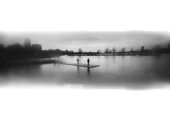 Two men stand on a dock extending into a wide body of water with a city skyline in the distance.