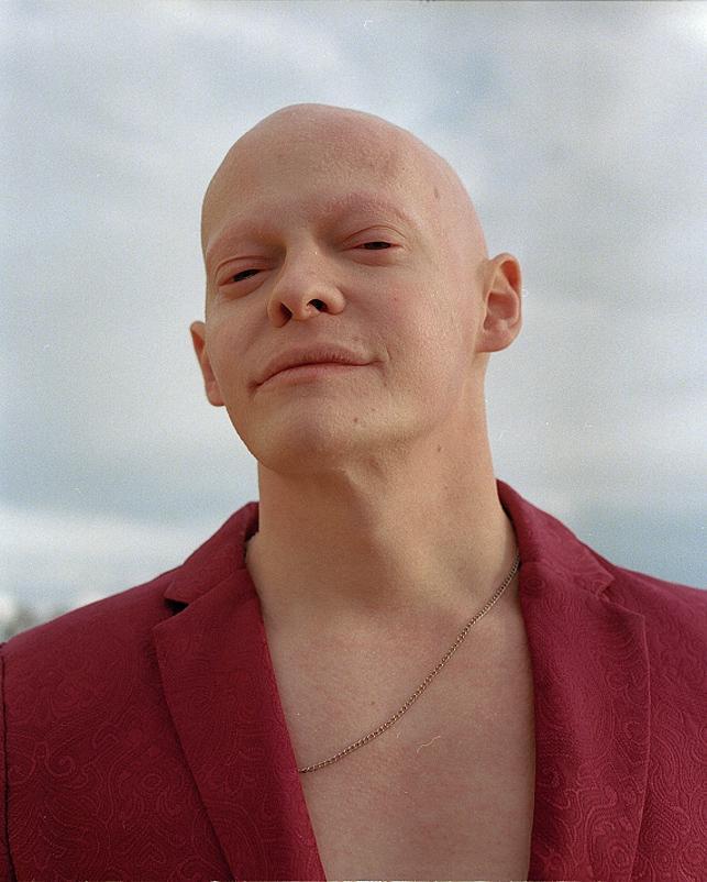 A man with a bald head and a red patterned jacket looks directly at the viewer.