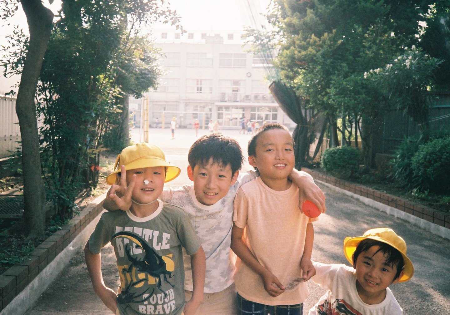 Four boys pose for a photo on a sunny path with a building and trees in the background.
