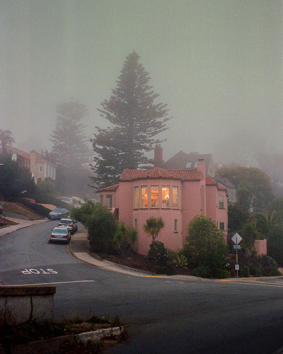 A pink house with illuminated windows sits on a street corner on a hazy day.